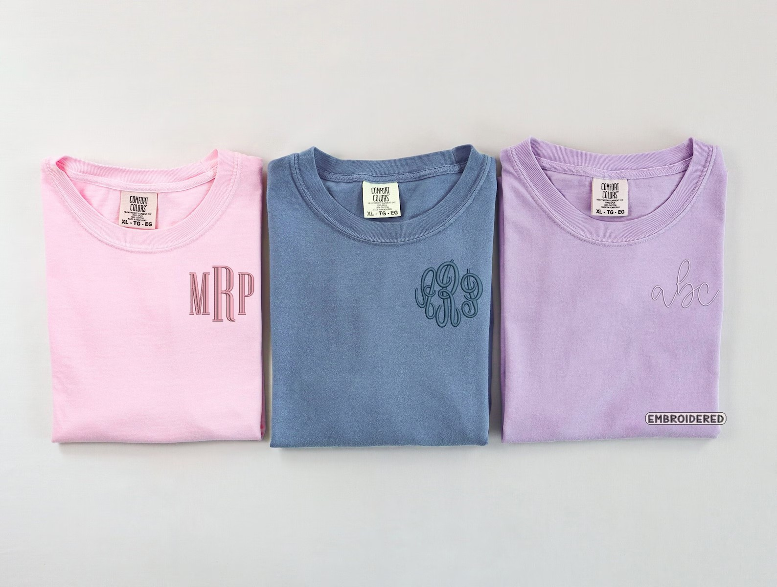 Three folded t-shirts in pink, blue, and purple with embroidered monograms on a light gray background.